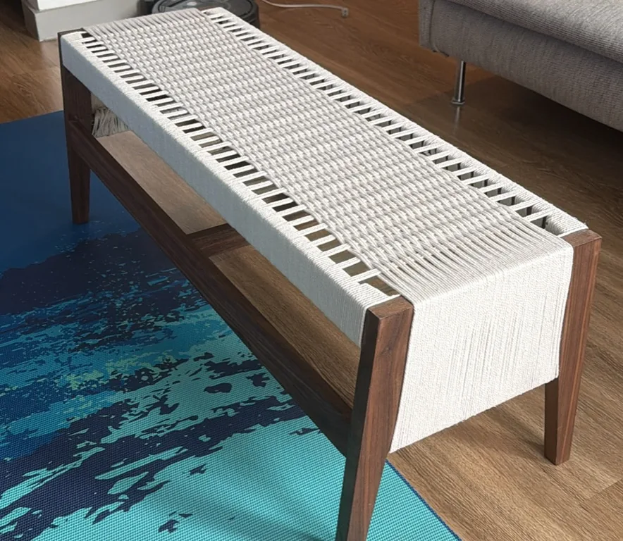 A long low bench seen from a raised three quarter angle, a dark walnut frame with splayed legs carrying a cream coloured hand woven rope seat, its fringe draping over the near end