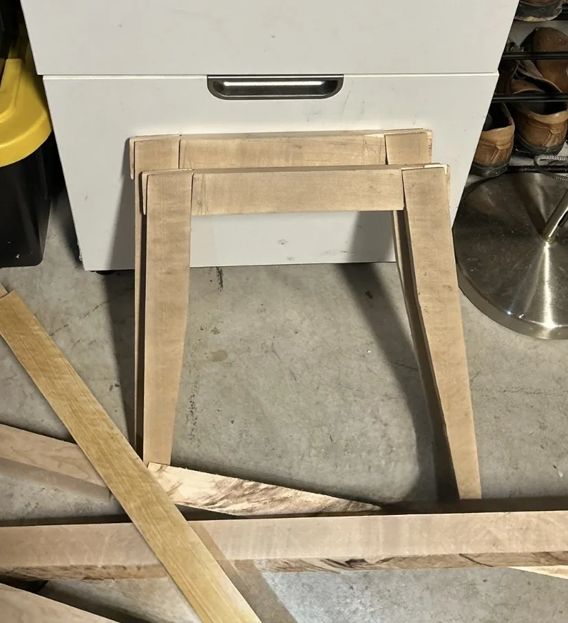 Two identical end frames in pale unfinished hard maple standing on a shop floor, each a pair of tapered legs joined by a top rail, with rail stock lying in front of them