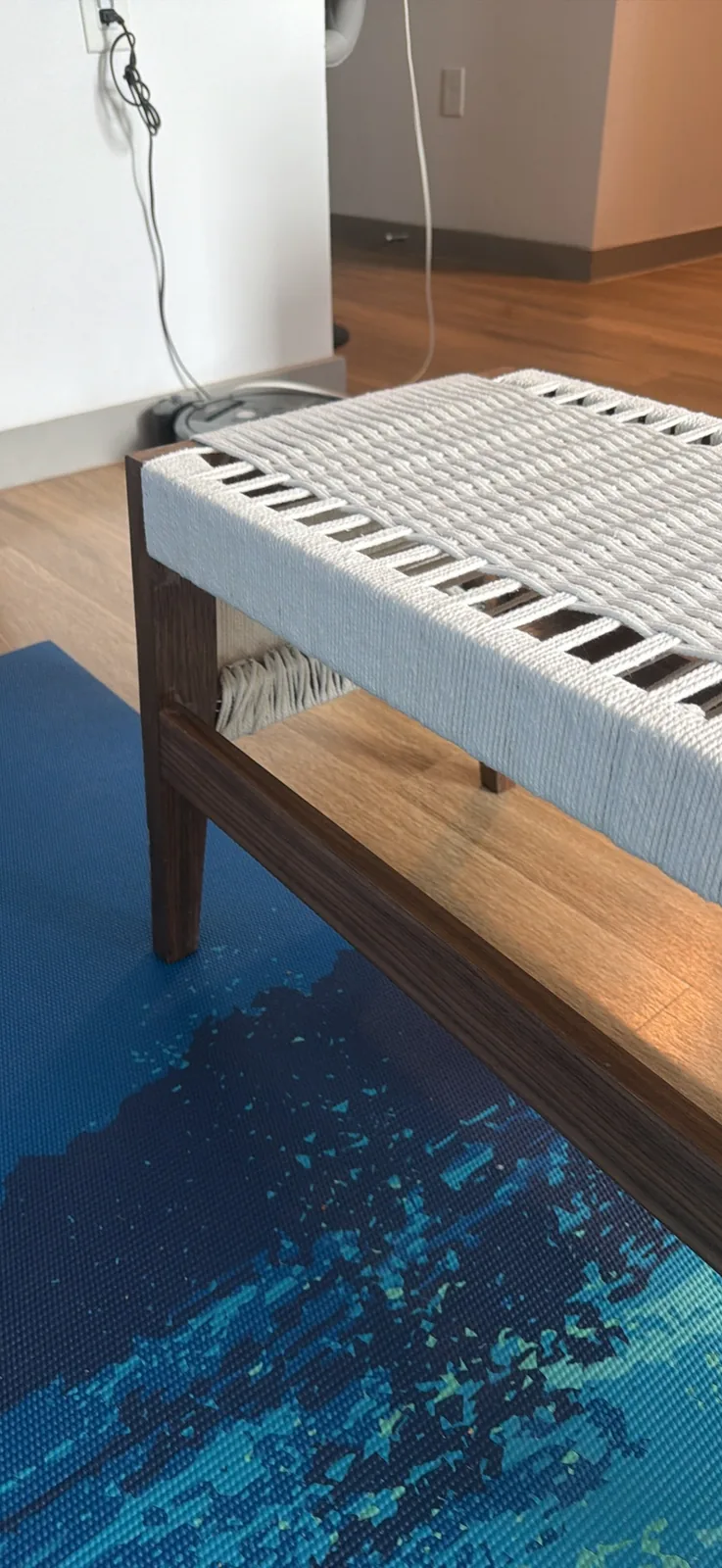 Close low view of one corner of the bench, showing a leg meeting the lower stretcher and the seat rail, with woven rope wrapping over the rail above
