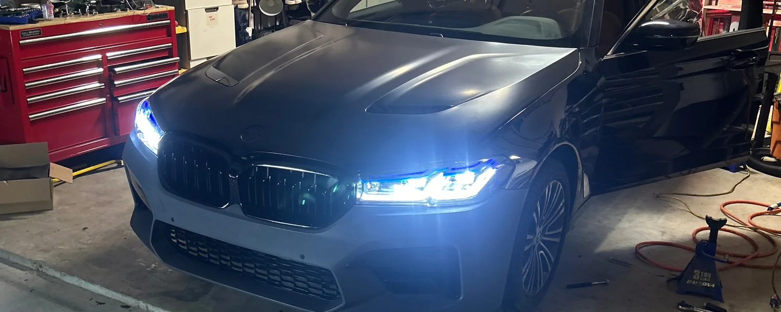 The BMW sedan in a home garage at night with its headlights on, seen from the front corner, with a red tool chest on the left and a jack stand and an air hose on the floor