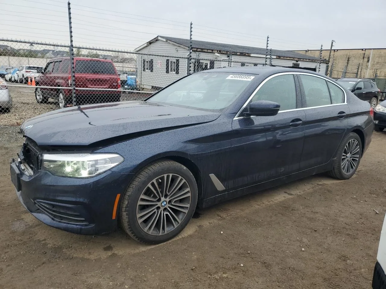 A dark blue BMW 5 Series sedan parked on dirt in a fenced salvage yard, seen from the front driver's side corner, with the bumper pushed in at the nose