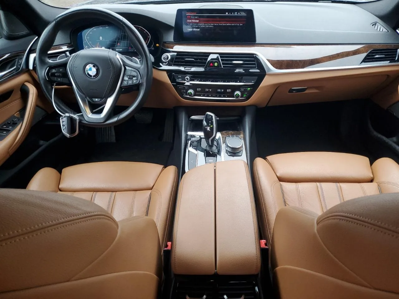 The front of the cabin seen from the back seat: cognac leather seats and armrest, a black dashboard with wood trim, the steering wheel and the centre screen