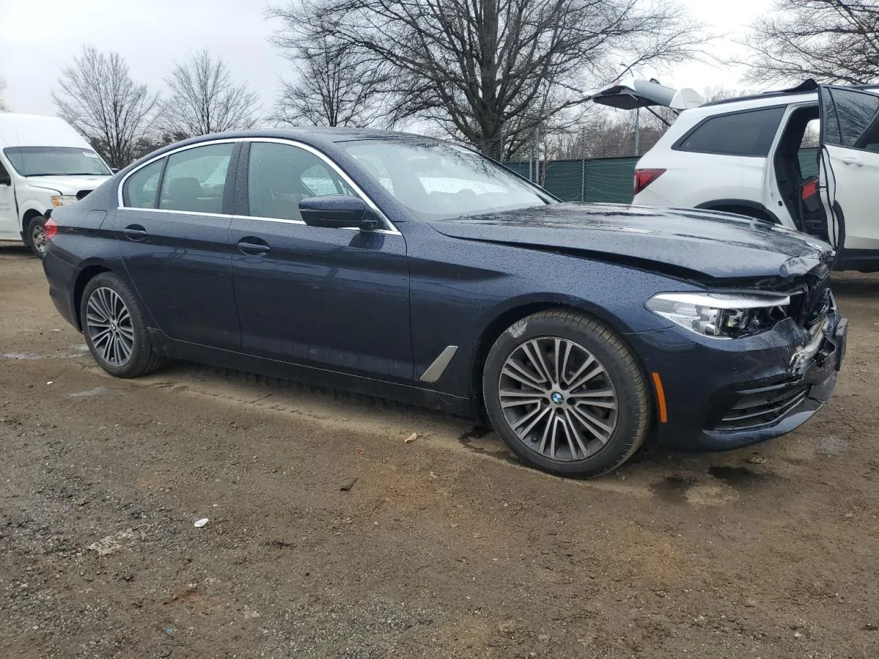 The dark blue sedan from its passenger side in the salvage yard, with the front bumper corner torn open and the headlight cracked