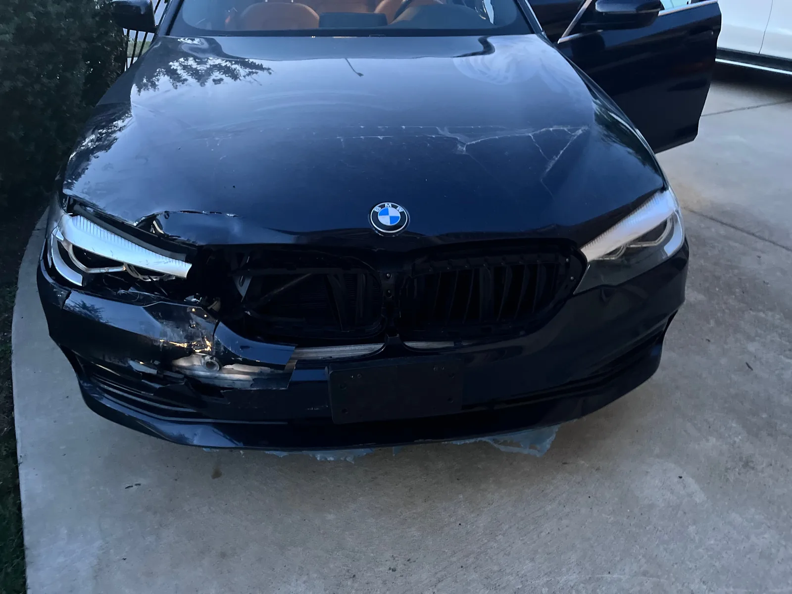 The front of the dark blue sedan on a concrete driveway, with a caved in grille, a split bumper and a broken headlight on one side