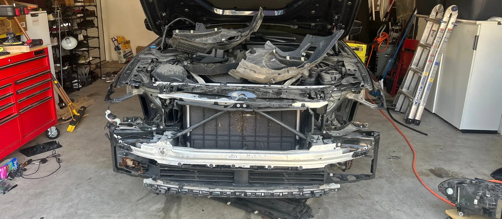 The front of the car in a garage with the hood open and the bumper cover, grille and headlights removed, showing the radiator, an aluminium bumper beam and loose plastic liners on top of the engine