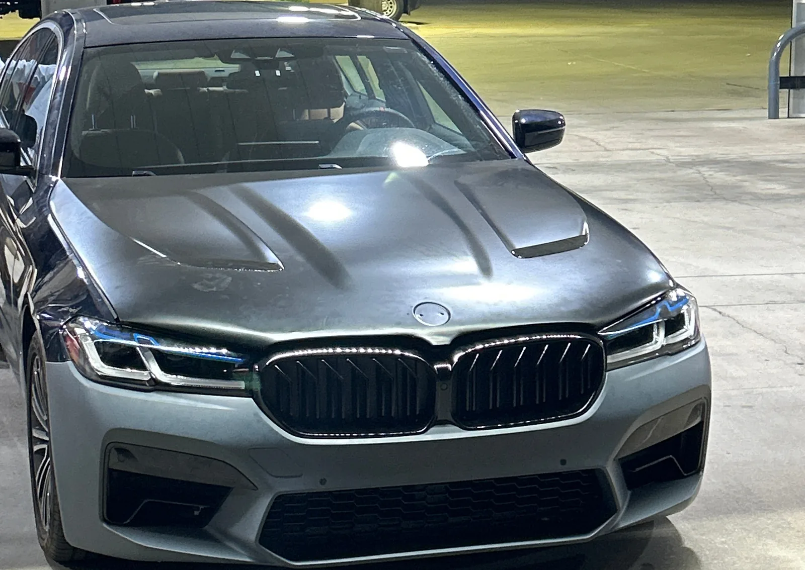 The sedan at a gas station at night, seen from the front, with a hood that has two raised bulges, a wide grille and a bumper with large intakes