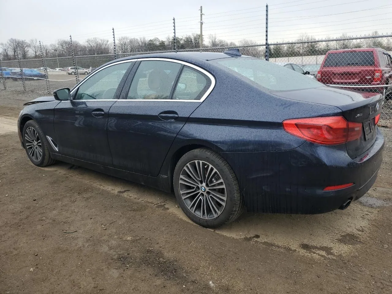 The dark blue sedan in a salvage yard seen from its rear corner on the driver's side, parked on wet dirt beside a chain link fence