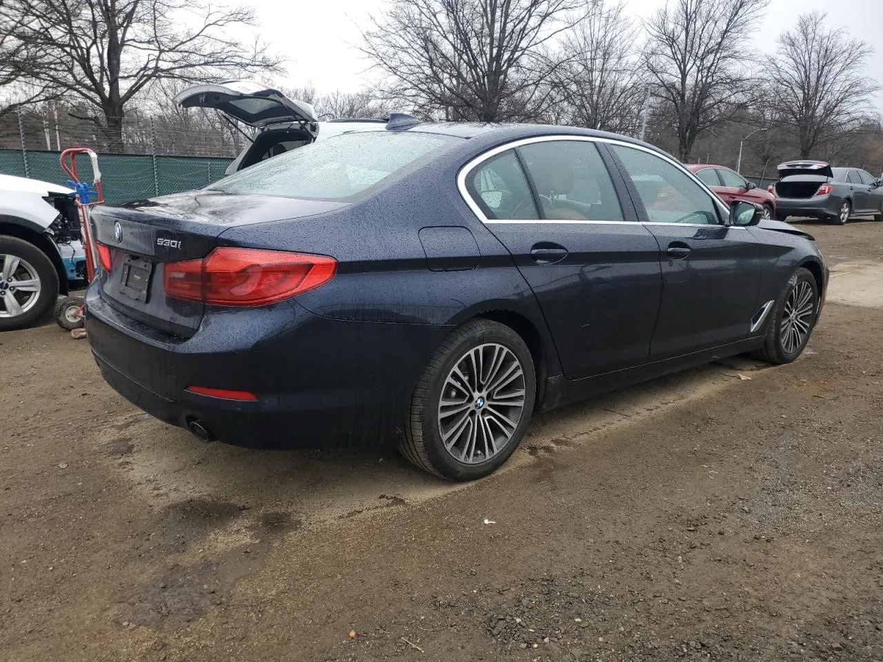 The dark blue sedan in a salvage yard seen from its rear corner on the passenger side, with other damaged cars and bare trees behind it