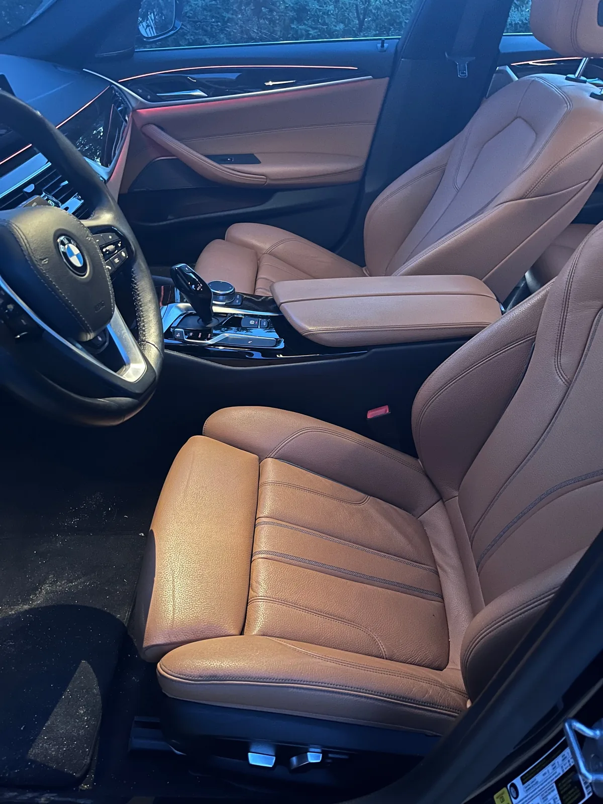 The cognac leather driver's and passenger seats seen through the open driver's door, with the steering wheel on the left and red light along the door trim