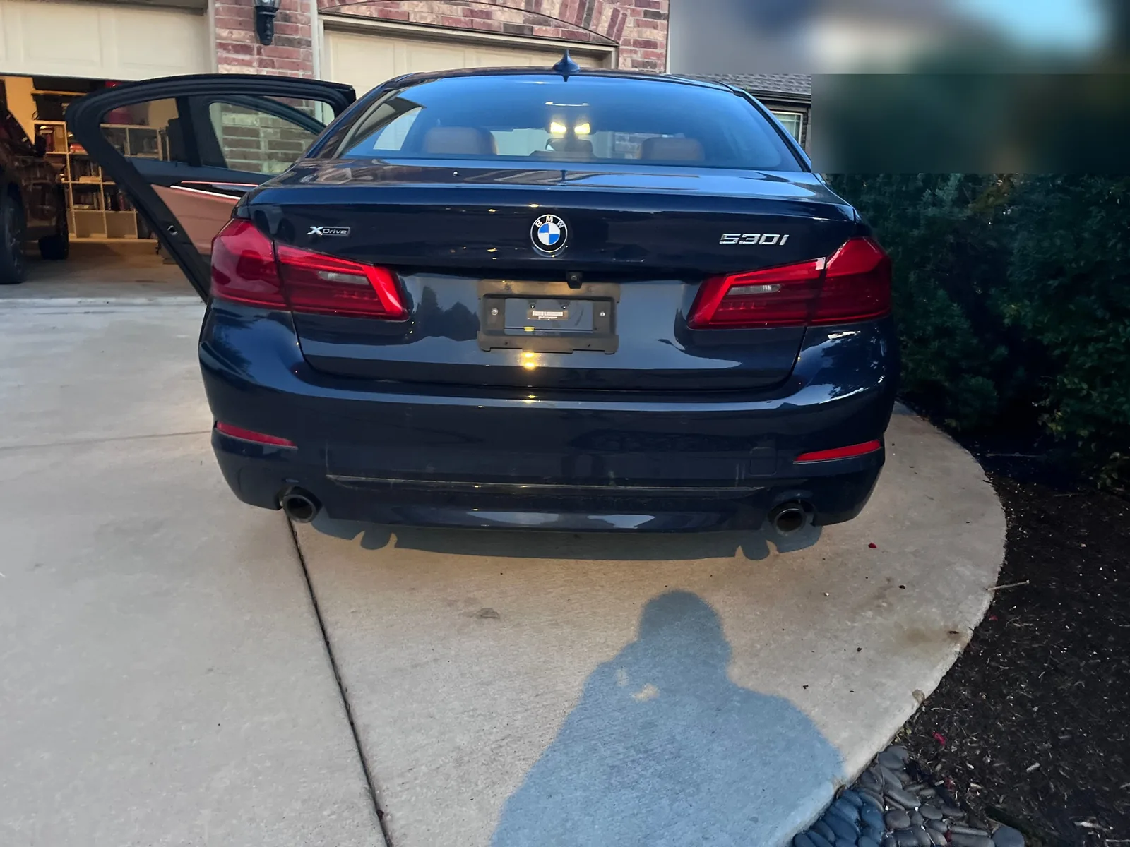 The rear of the dark blue sedan on a driveway at dusk, with xDrive and 530i badges on the trunk, an empty plate holder and a rear door open