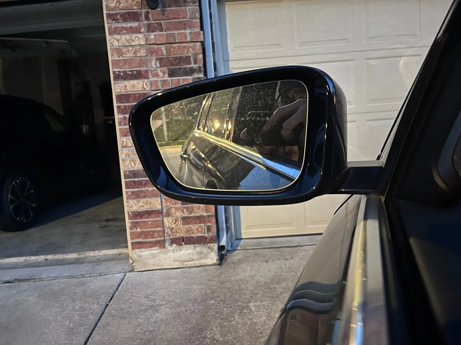 A dark blue side mirror on the car in front of a brick garage pillar, with the side of the car reflected in the dusty glass