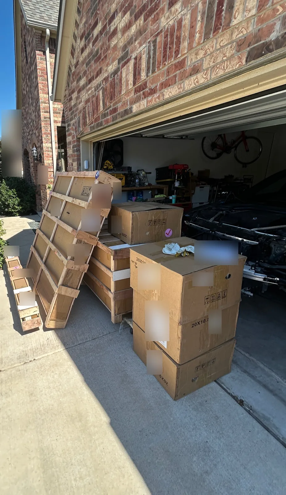 Wooden crates and stacked cardboard boxes of car parts on a driveway in front of an open garage, with the front of the car inside