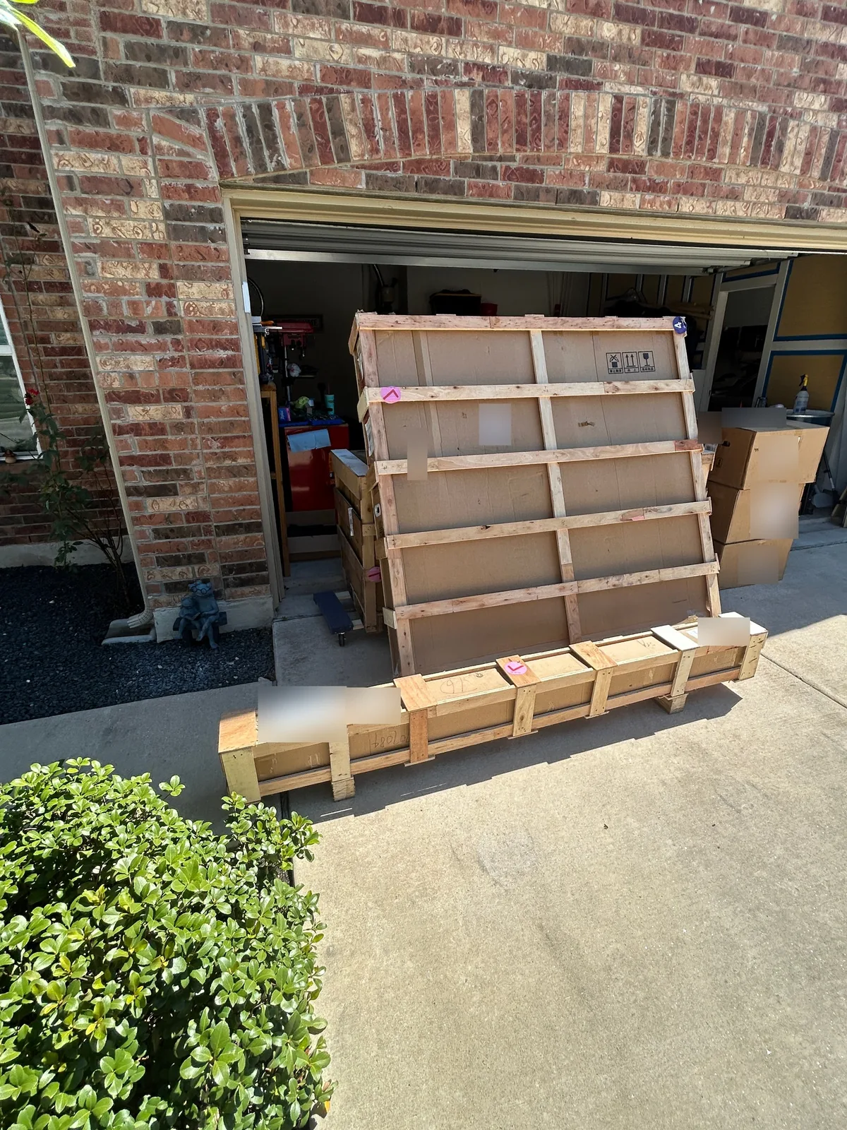 A tall flat wooden crate standing in an open garage door and a long narrow crate lying on the driveway in front of it, with more boxes stacked to the right