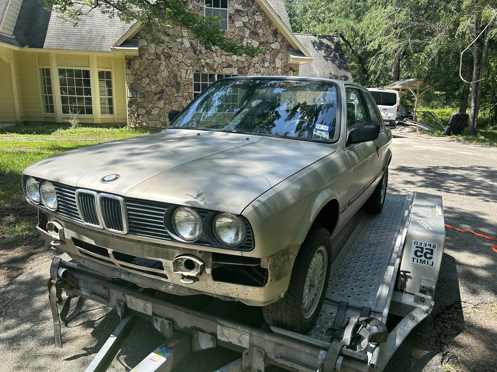 A champagne coloured 1984 BMW E30 two door strapped to a U-Haul car trailer on a driveway, seen from the front three quarter, with trees and a house behind it