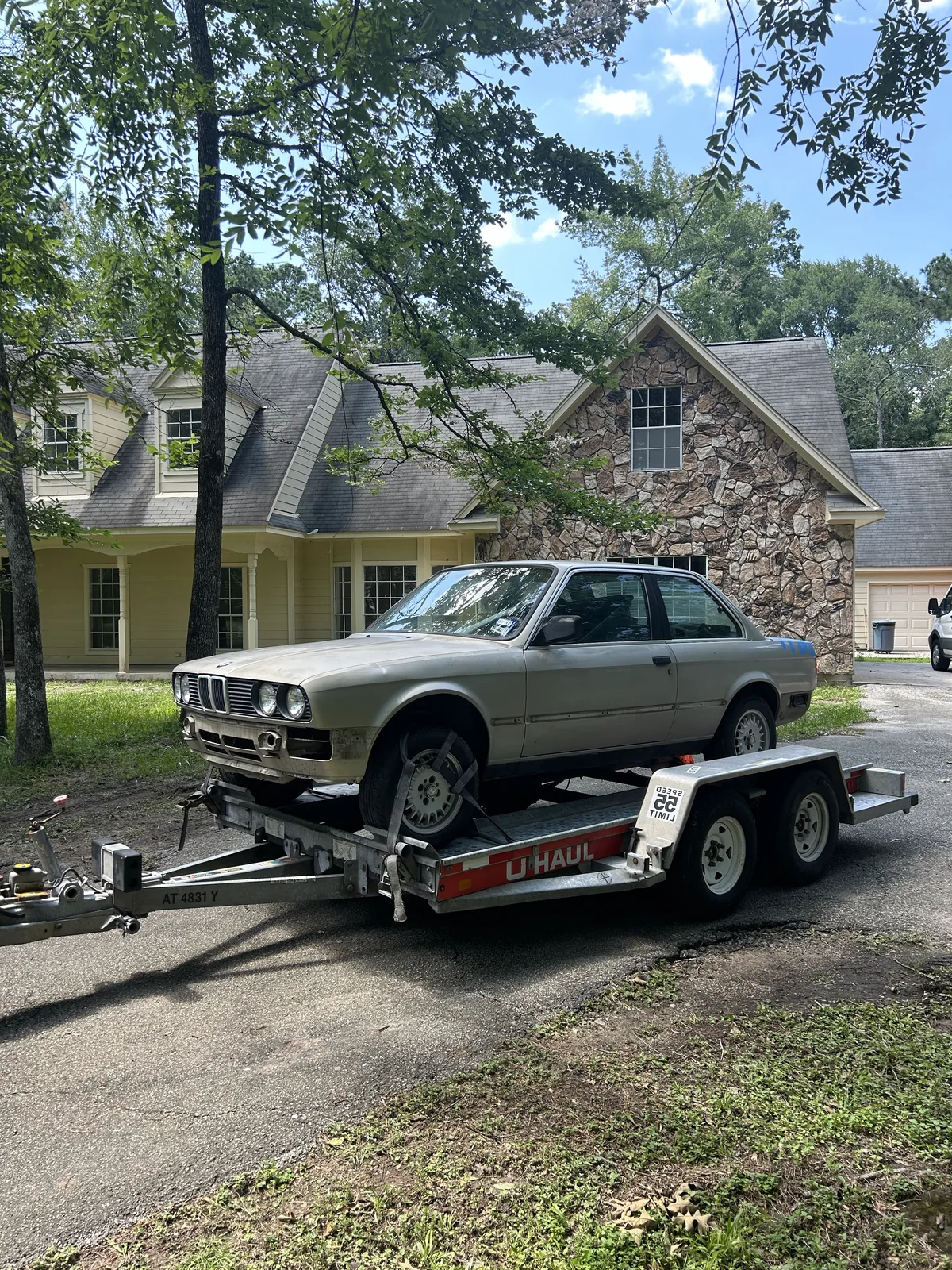 The E30 on its trailer in front of a stone and stucco house, seen from the front three quarter in bright daylight
