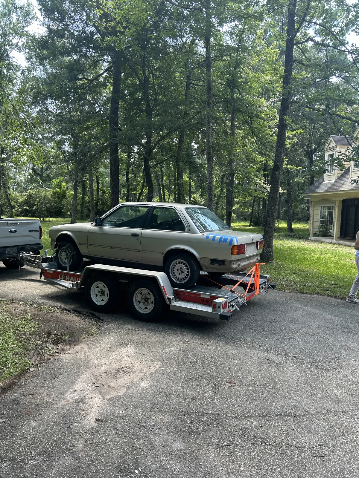 The E30 sitting on the trailer in a wooded driveway, viewed from the side, with blue tape on the rear quarter panel