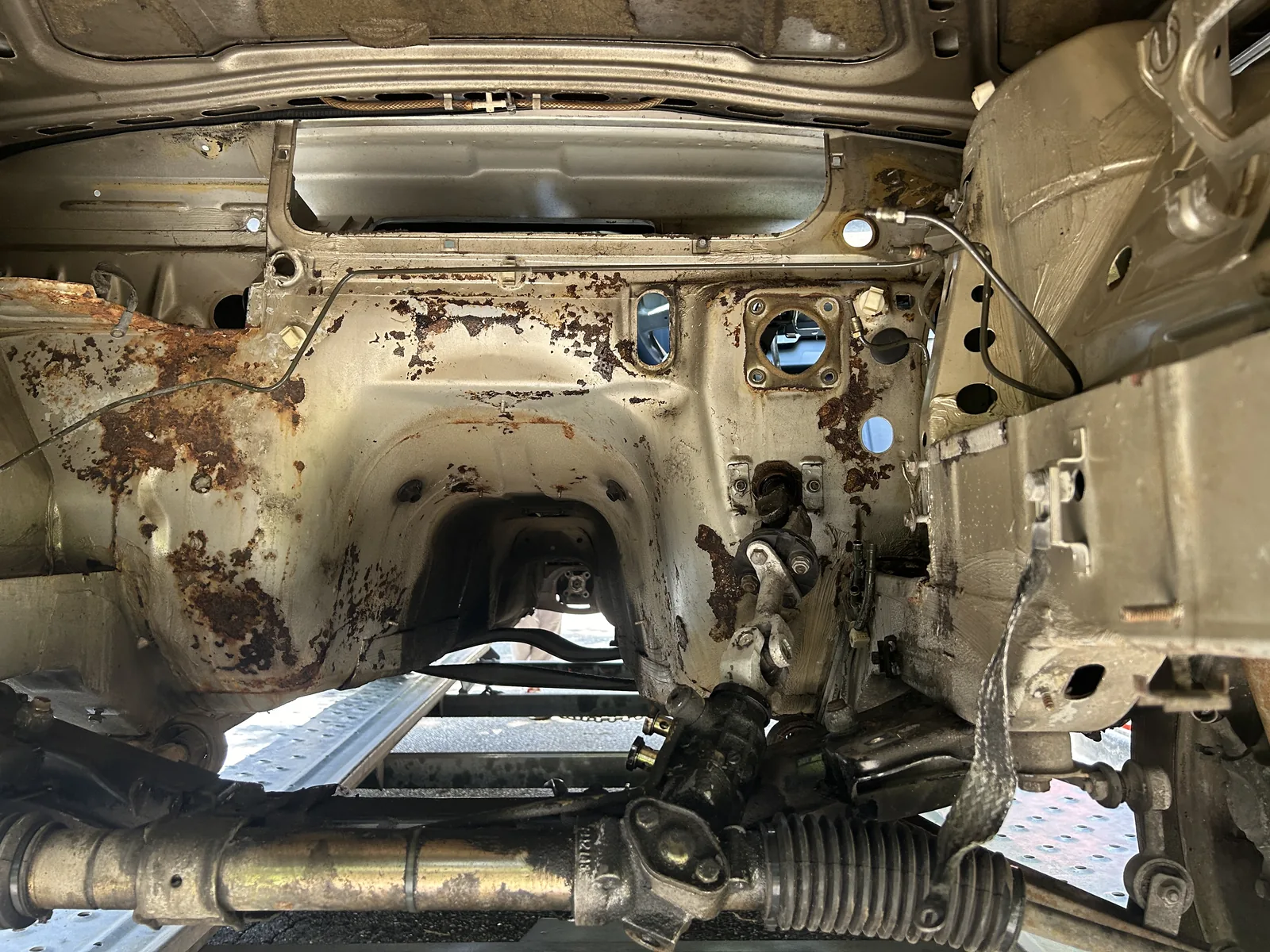 The empty engine bay of the E30 with the engine already removed, showing bare metal, surface rust across the inner wings and the steering rack still in place