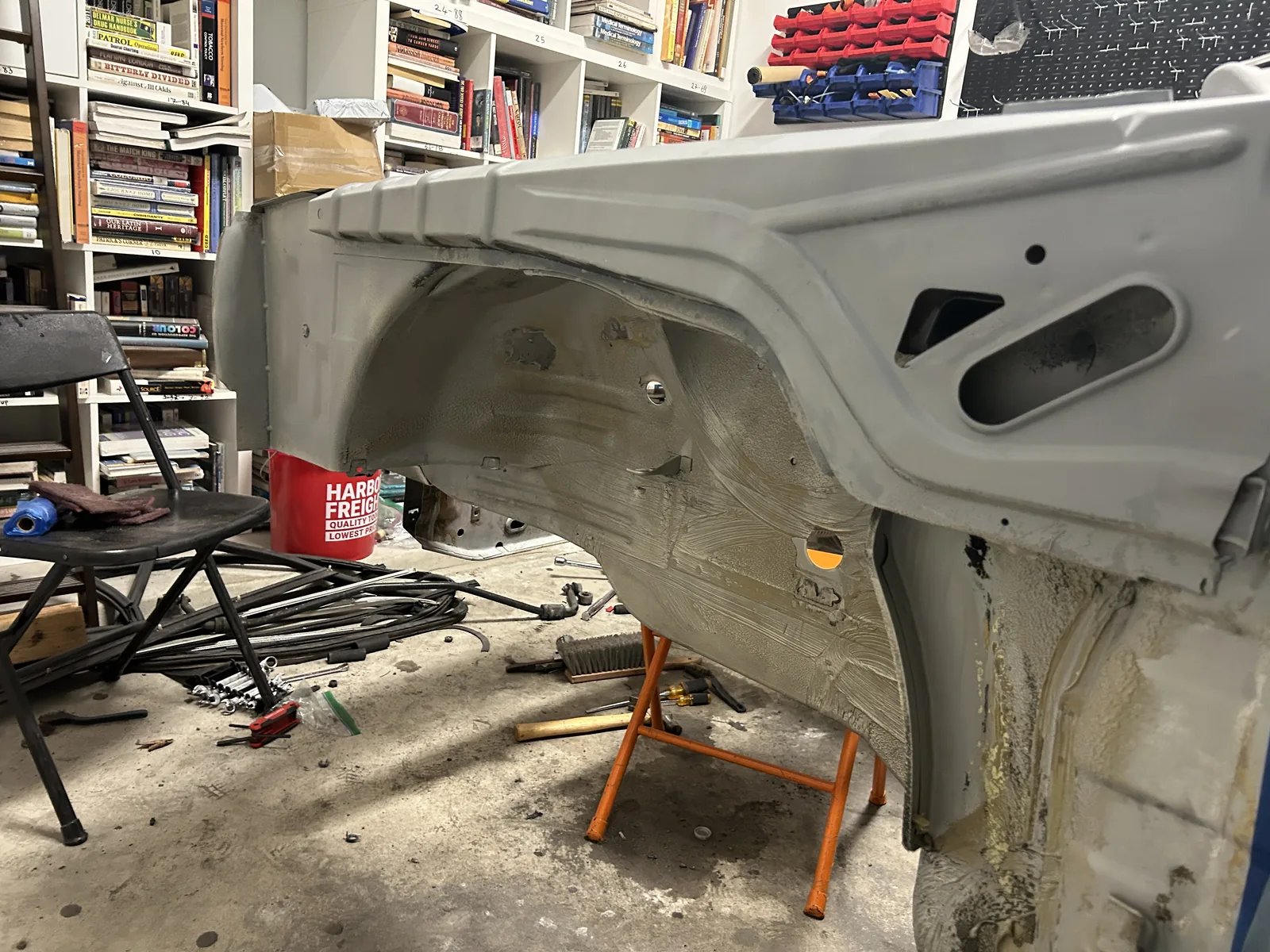 The removed front end panel of the E30 standing on a shop floor next to the car, with shelving and tools in the background