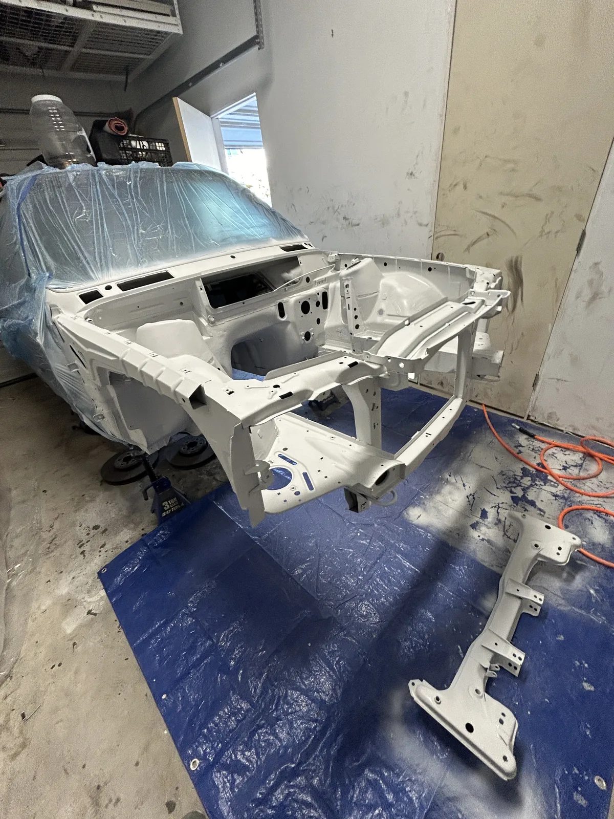The primed E30 shell in a workshop with its front structure and crossmember laid out separately on a blue tarpaulin on the floor