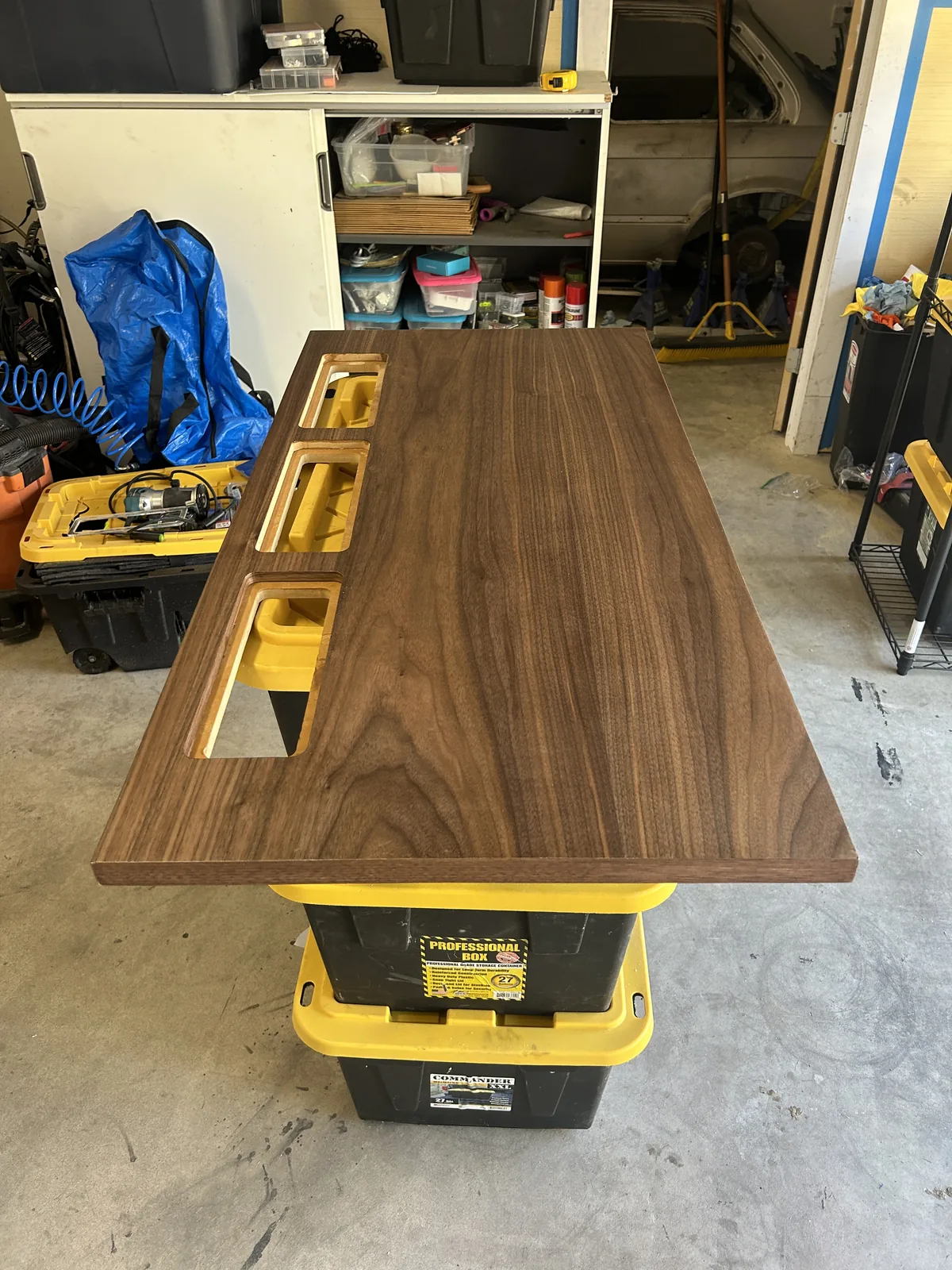 A walnut desk top resting on storage totes in a garage, with three identical rectangular ports cut along one edge