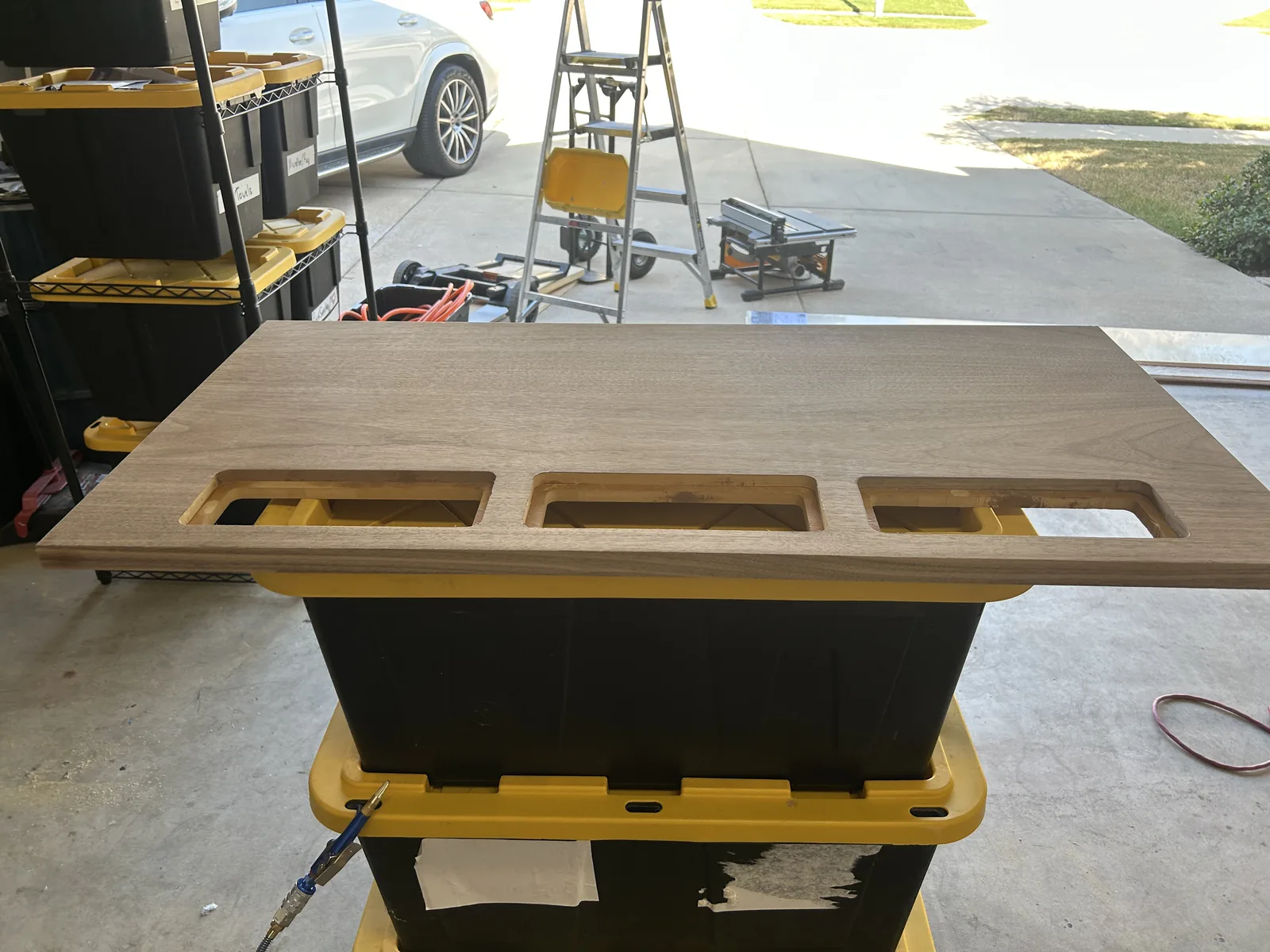The walnut desk top laid across two plastic storage totes in an open garage, with a driveway and ladder visible behind