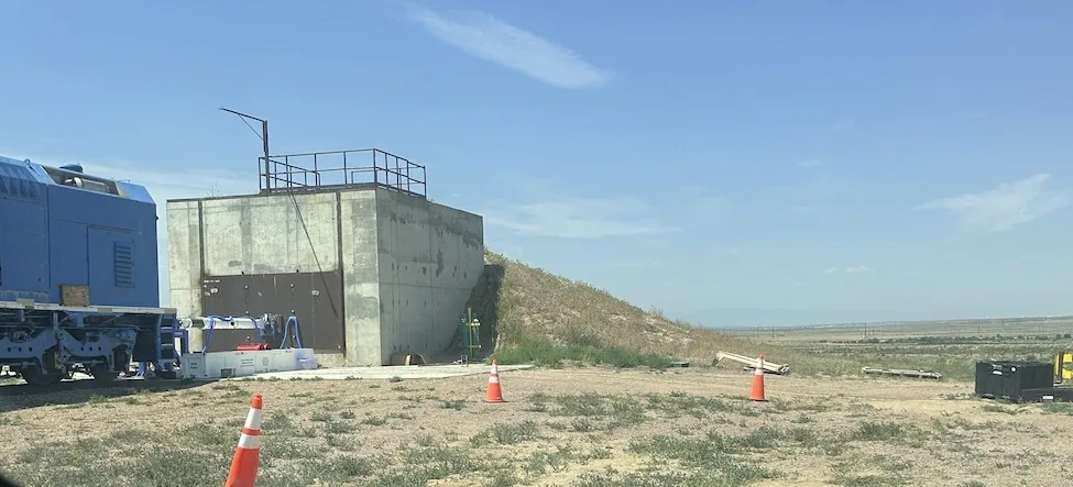 A reinforced concrete test cell built into an earth berm under a wide blue sky, with a blue locomotive on rails to the left, safety cones in the foreground and open high desert stretching to the horizon
