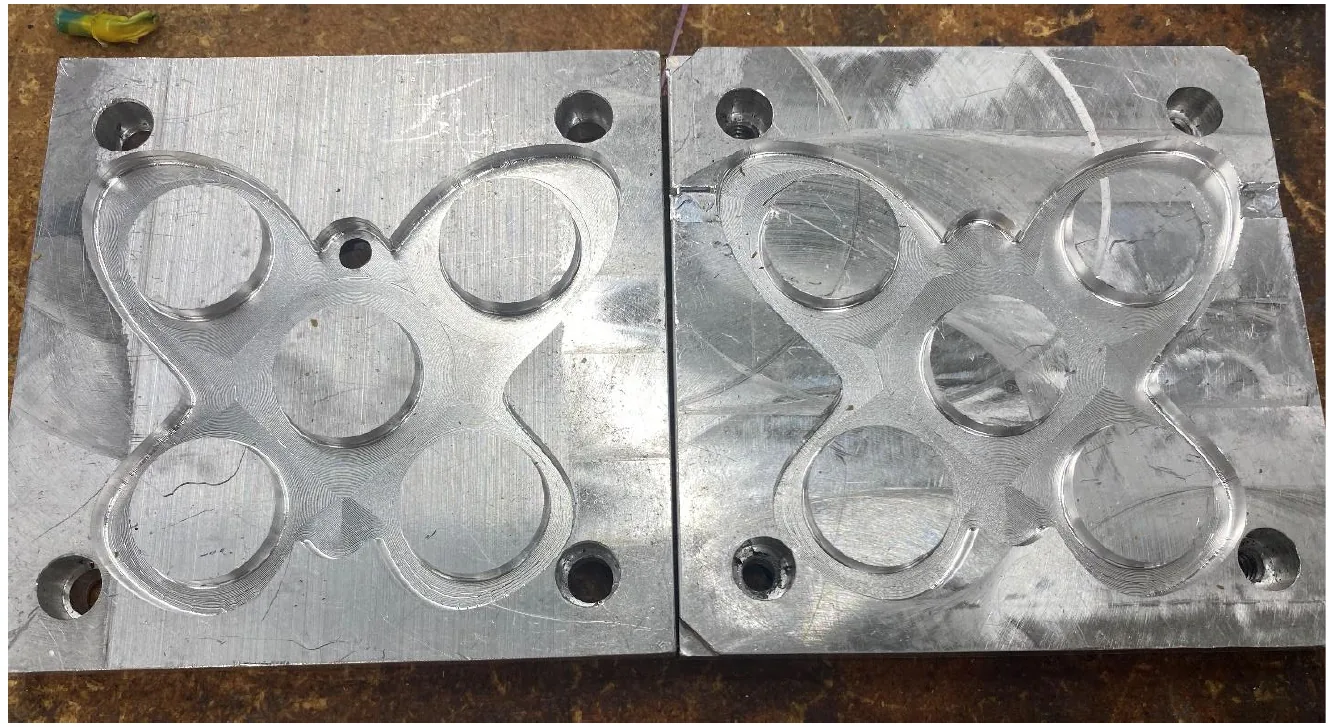 Two machined aluminium blocks side by side on a workbench, each with a butterfly shaped cavity milled into it