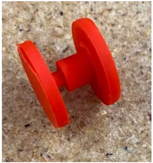 Two red 3D printed discs joined by a short shaft, resting on a concrete surface