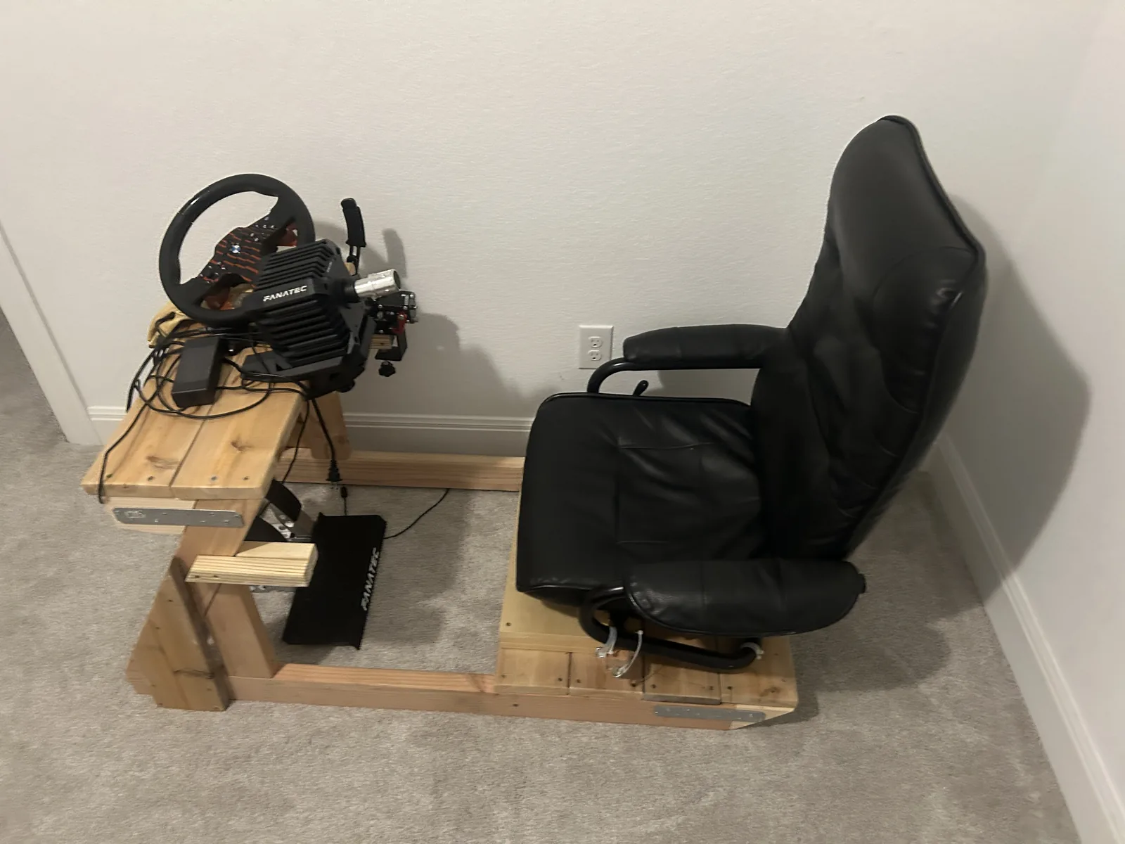 A sim racing rig built from dimensional timber, with a direct drive wheel base on a raised deck, pedals ahead of it and a black bucket seat behind