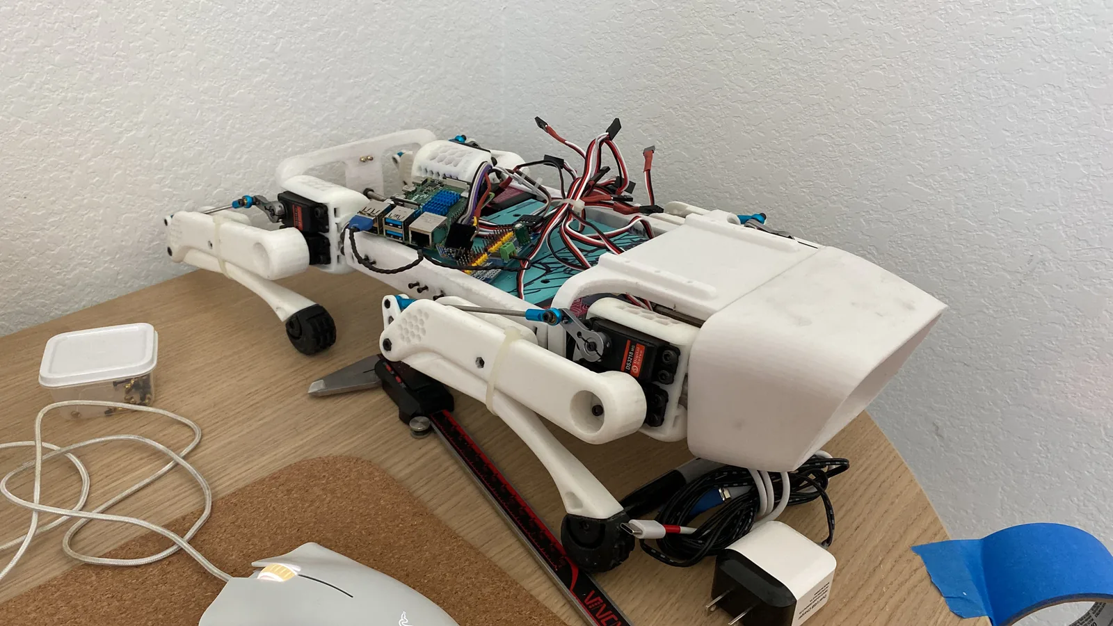 A white 3D printed four-legged robot standing on a desk, its body open with wiring visible