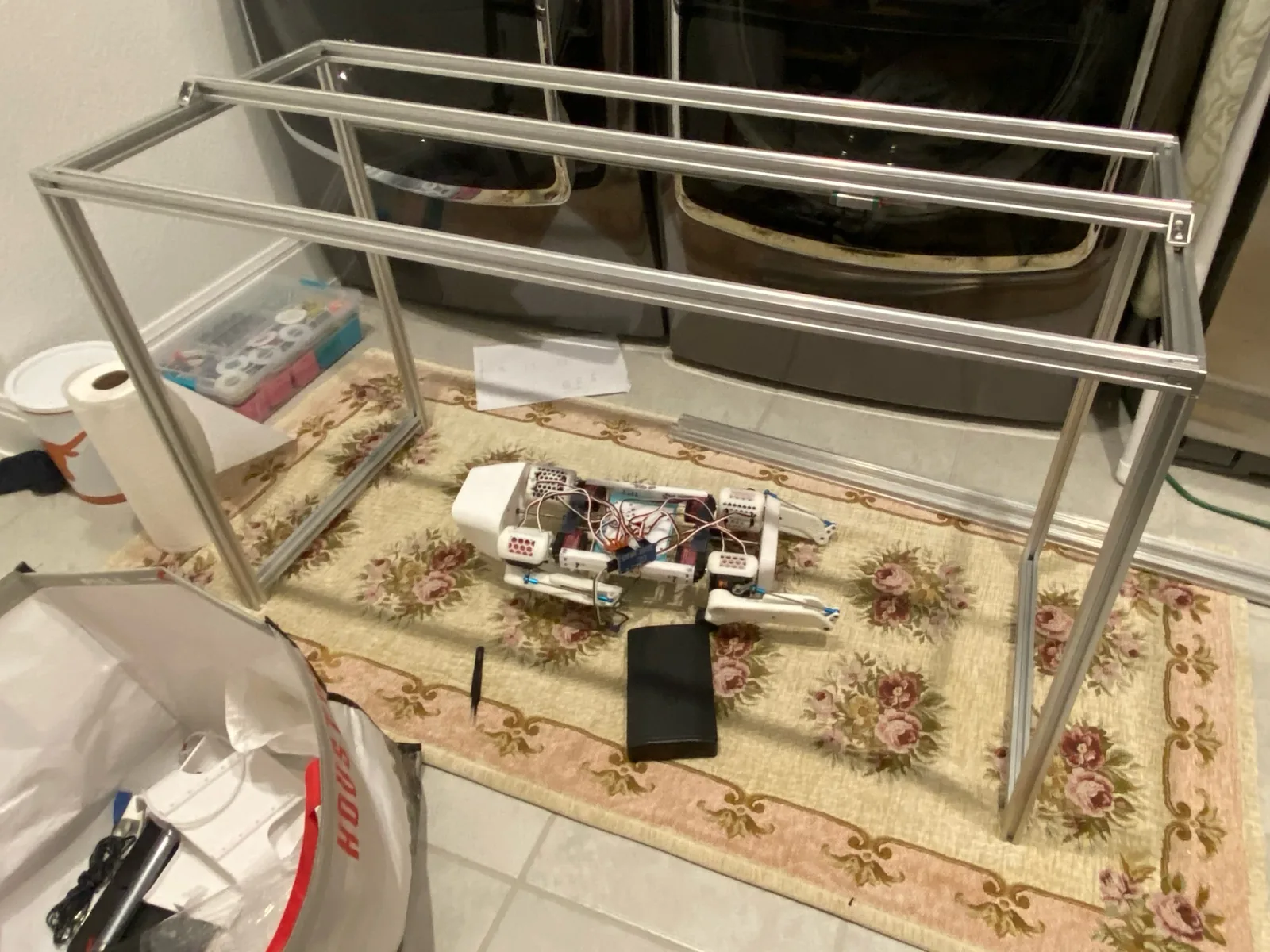A white four-legged robot with its cover off lying on a floral rug beside a rectangular aluminium extrusion frame