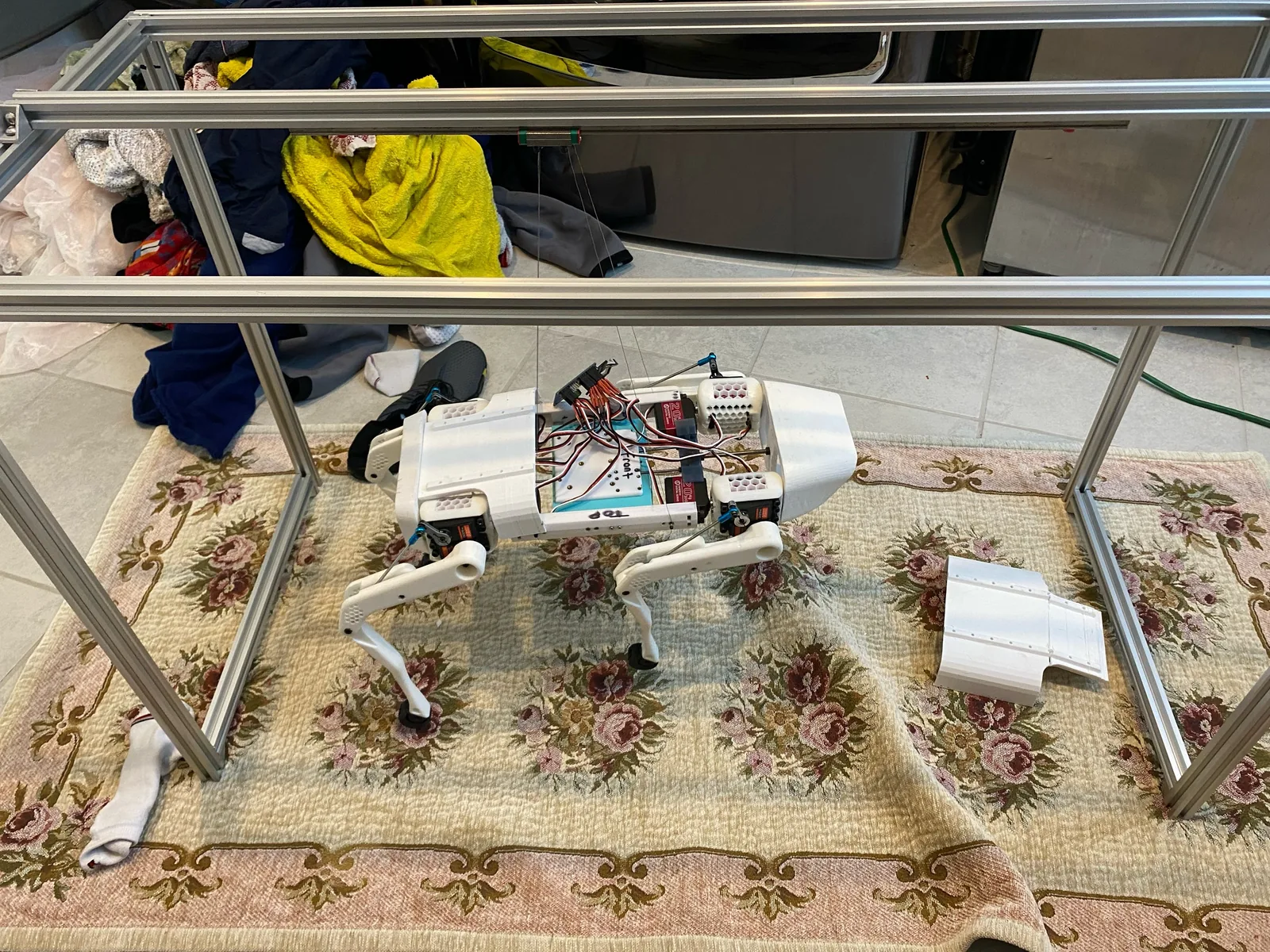 A white four-legged robot with its cover off standing on a floral rug inside an aluminium extrusion frame, thin lines running from the top rail to its body