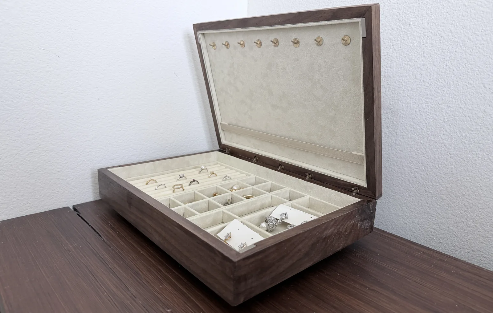 A dark hardwood jewelry box open on a dark wood desk against a white wall, its cream lined lid raised and a compartment tray of rings and earrings filling the base