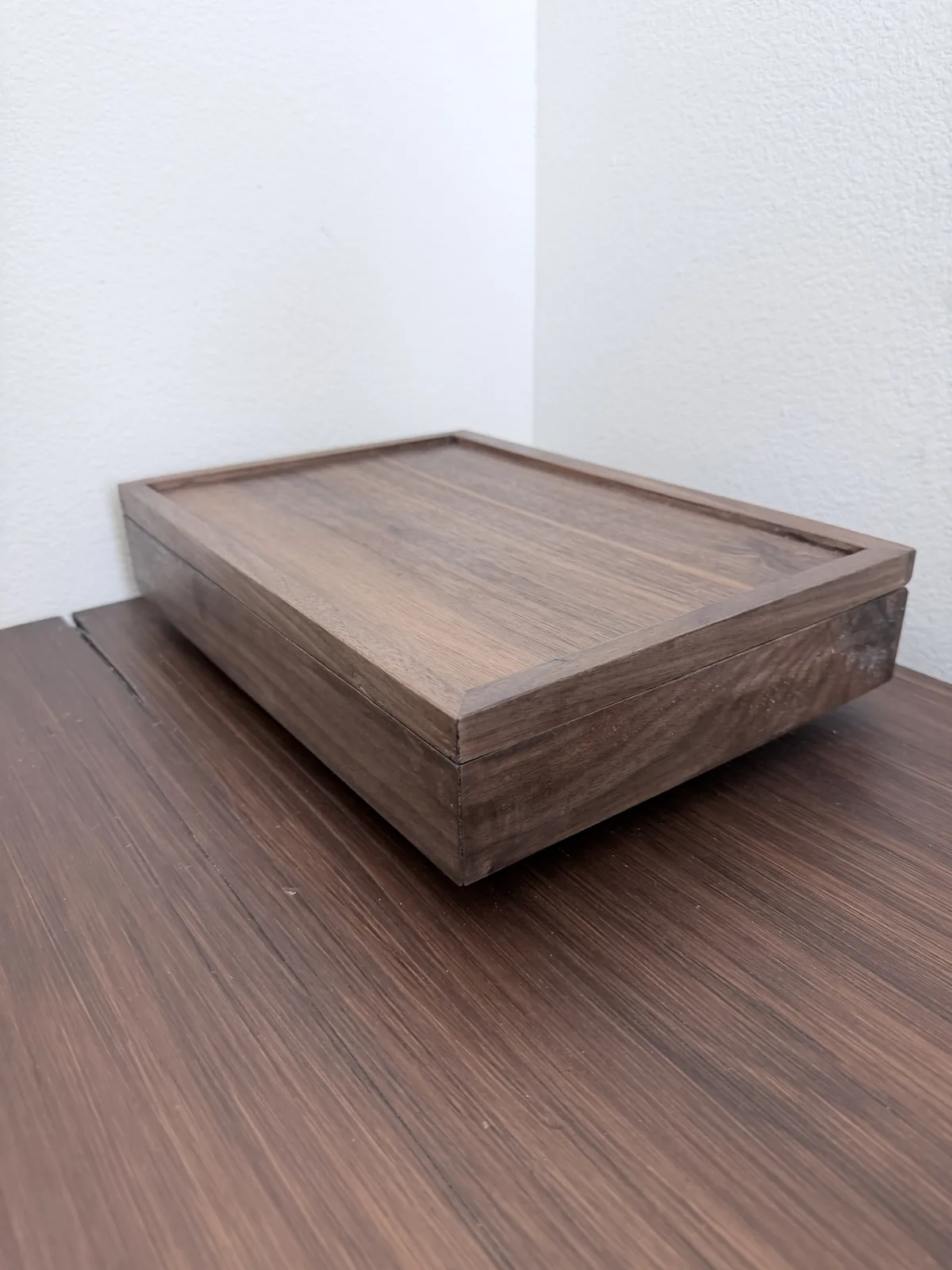 The closed jewelry box on a dark wood desk against a white wall, seen from a raised three quarter angle, showing the mitred lid frame and the grain of the panel