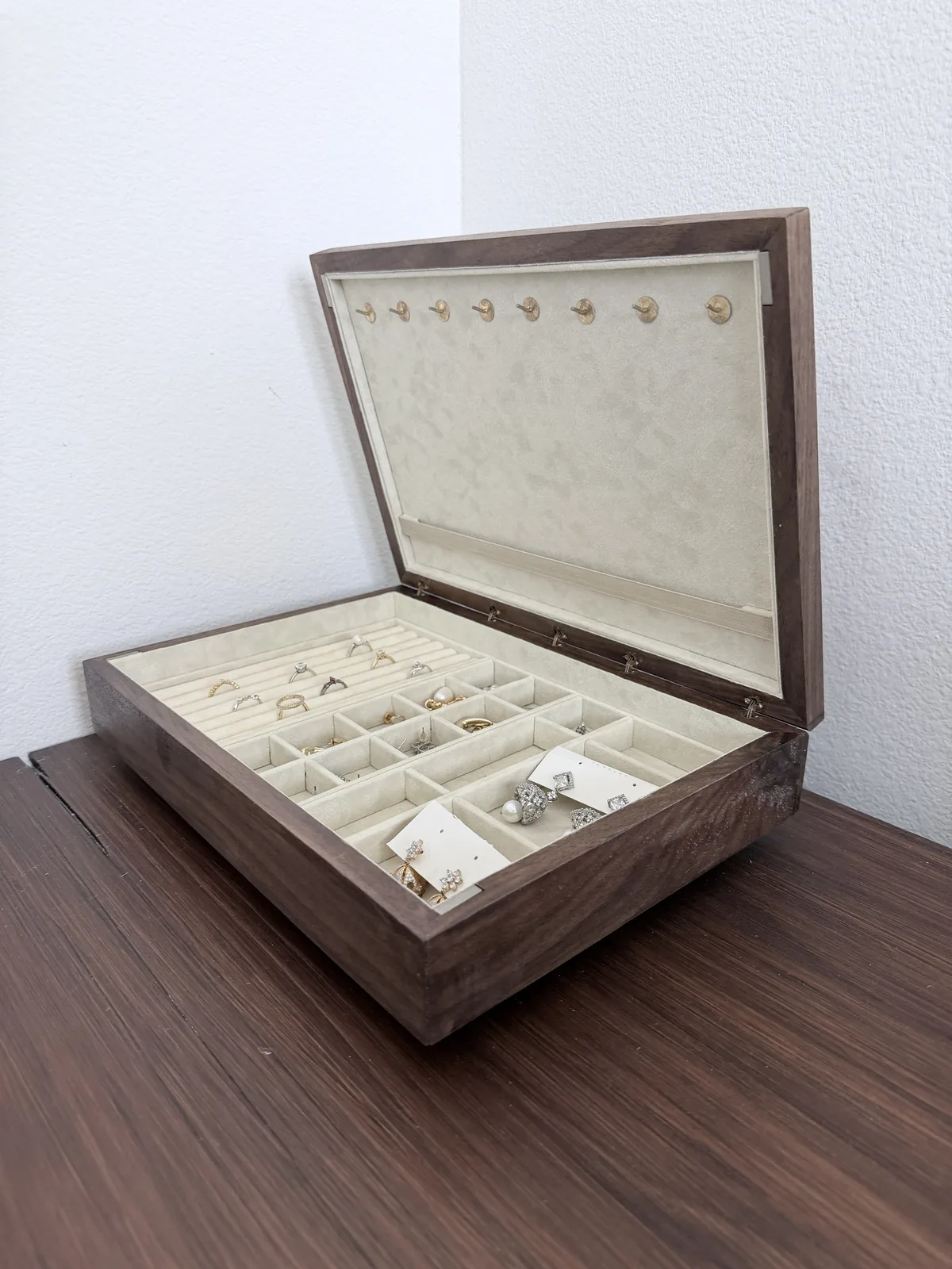 The jewelry box open on a dark wood desk, photographed from the front, the lid standing open against a white wall and the loaded tray filling the base