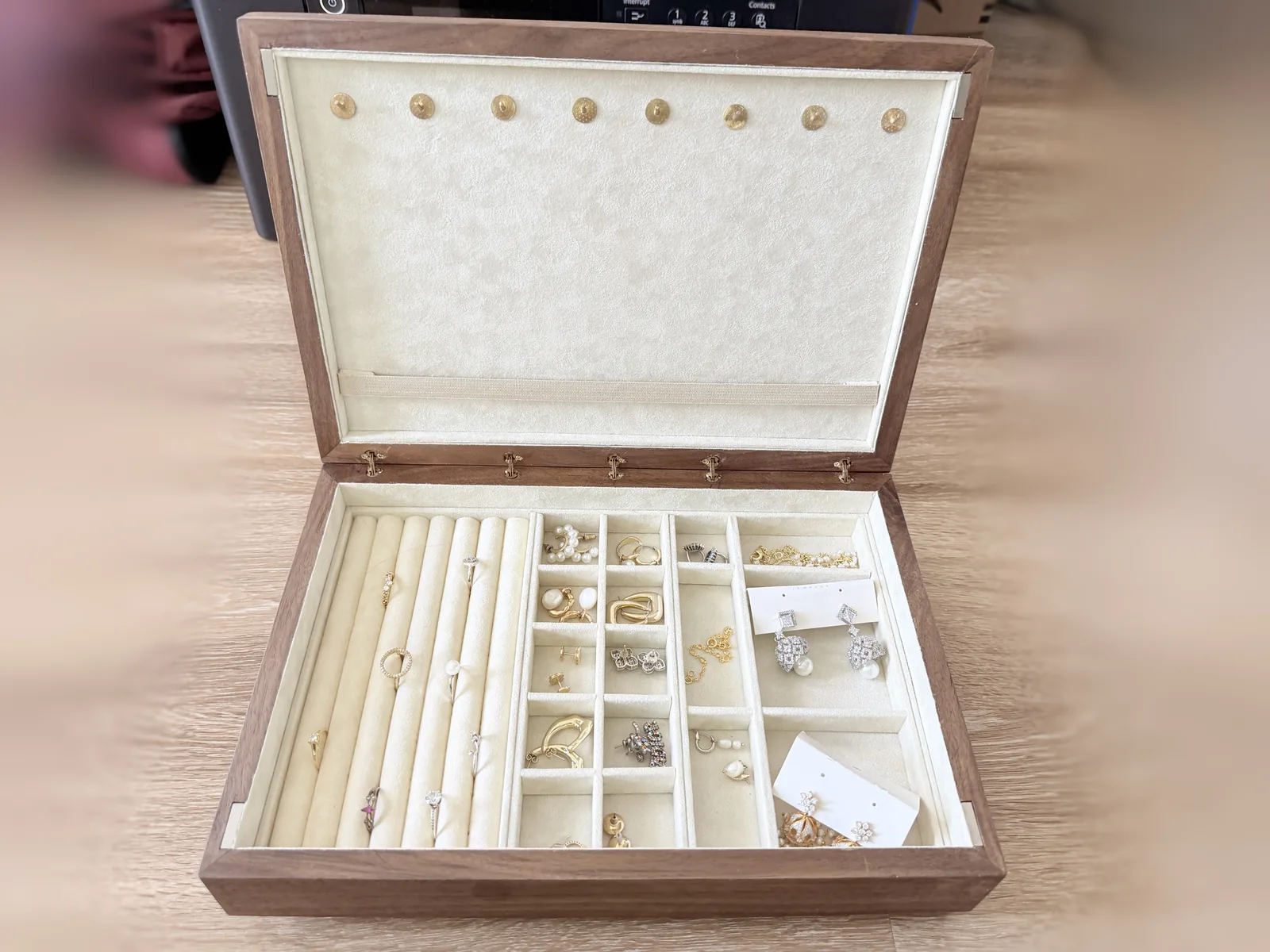A dark hardwood jewelry box open on a pale wood floor, lined in cream, a compartment tray full of rings and earrings filling the base and a row of small brass hooks along the top of the lid lining
