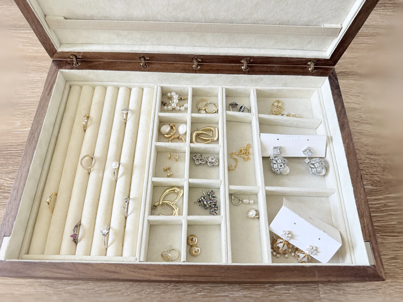 Looking down into the open jewelry box at the cream lined tray, padded ring rolls holding rings on the left and a grid of small compartments holding earrings on the right