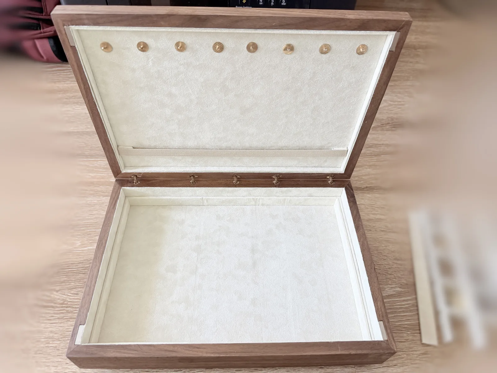 The jewelry box open and empty on a wood floor, its cream lined interior bare, with the removable compartment tray sitting to the right of it