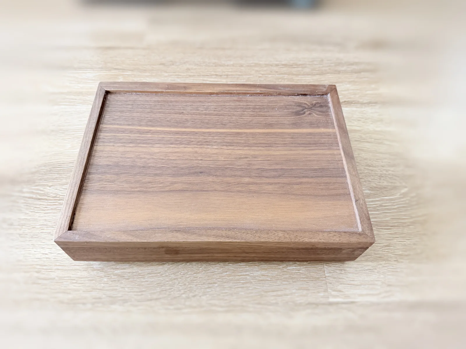 The closed jewelry box resting on a pale wood floor, its lid a mitred frame surrounding a panel of horizontal grain