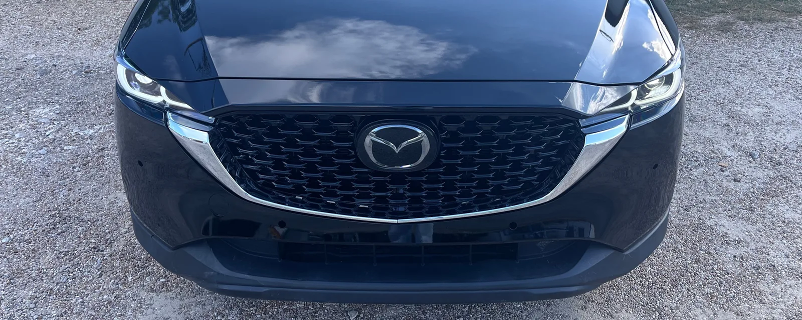 The front of a black Mazda CX-5 seen head on, with a glossy hood reflecting clouds, a black mesh grille with chrome trim and the Mazda badge, parked on gravel