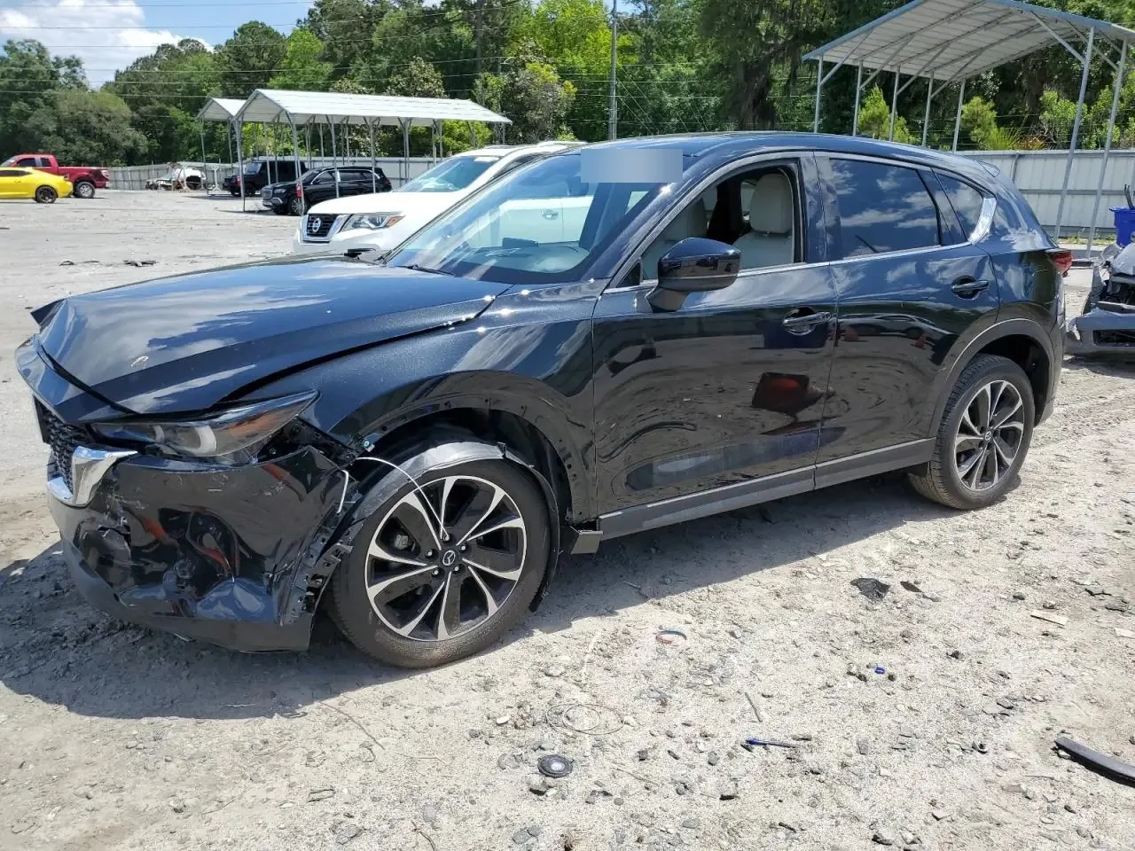 A black Mazda CX-5 on a dirt auction lot seen from the front left, with the bumper torn open at the corner and the hood buckled. The lot sticker on the windshield is blurred
