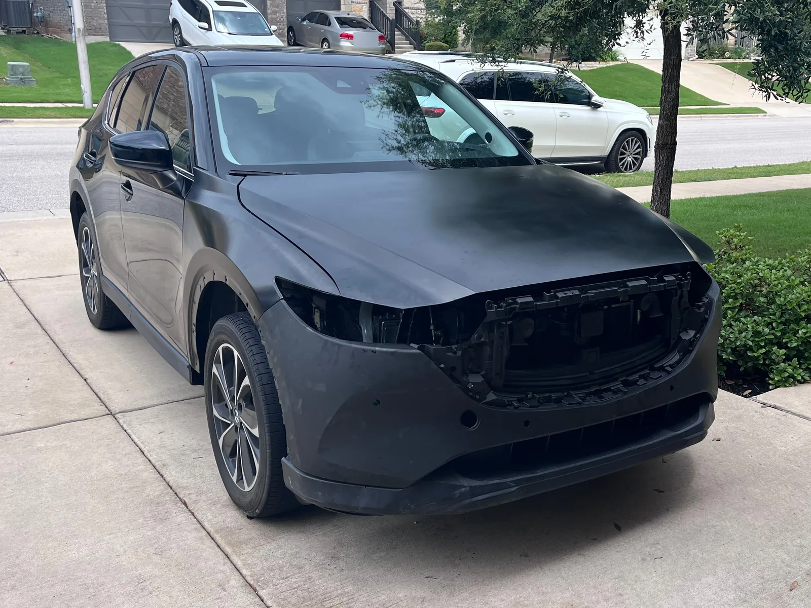 The CX-5 in daylight from the front left, with a dull grey hood and bumper cover and an empty grille frame where the grille goes
