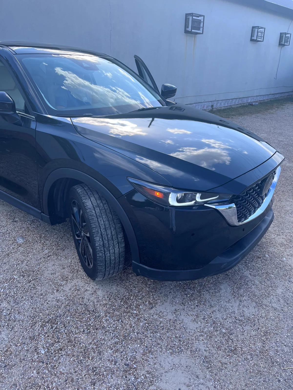 The finished CX-5 from the front right, glossy black with the grille and headlight in, parked on gravel beside a grey building