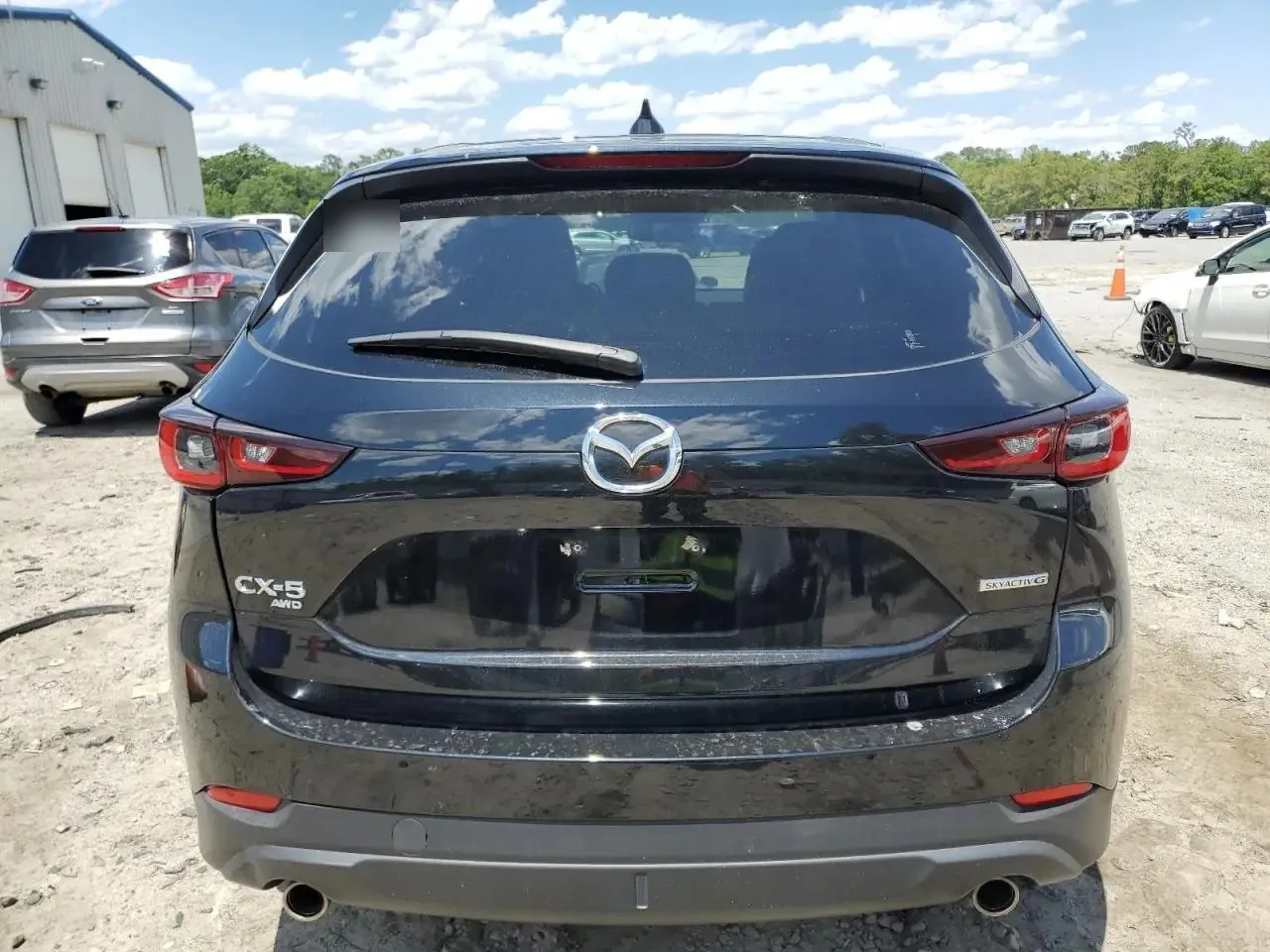 The rear of the black CX-5 on the dirt auction lot, with the Mazda badge, the CX-5 AWD and Skyactiv-G badges on the tailgate and twin exhaust tips. Marker writing on the rear glass is blurred