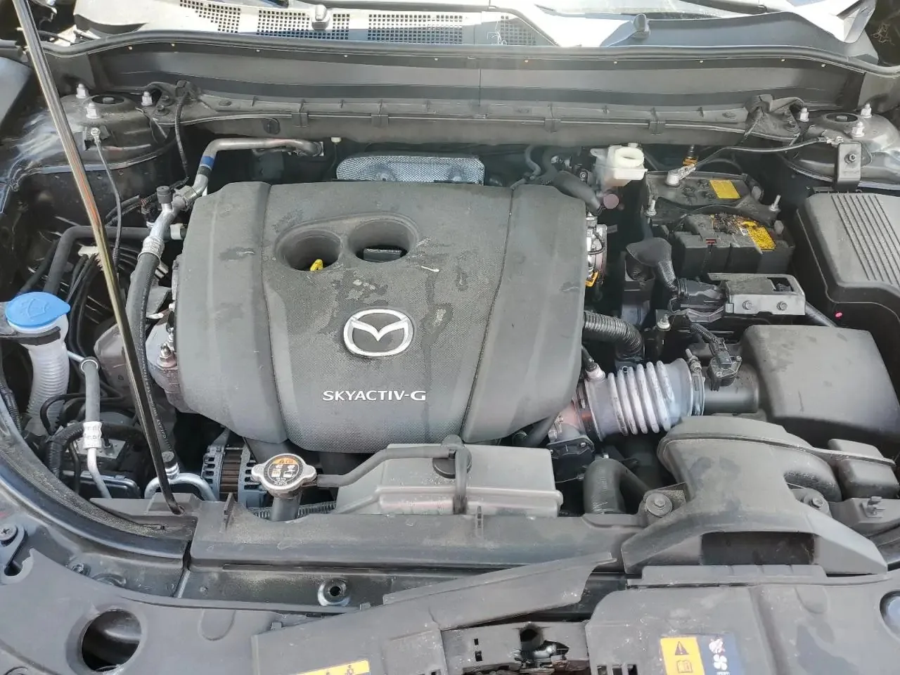The CX-5's engine bay with the hood propped open, showing the black engine cover marked Skyactiv-G, the battery and the washer fluid cap