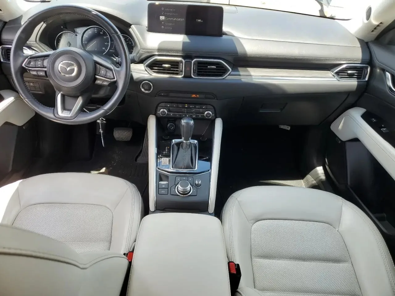 The CX-5's dashboard seen from behind the front seats, with the steering wheel, the screen on top of the dash, the gear selector and the two light grey seats