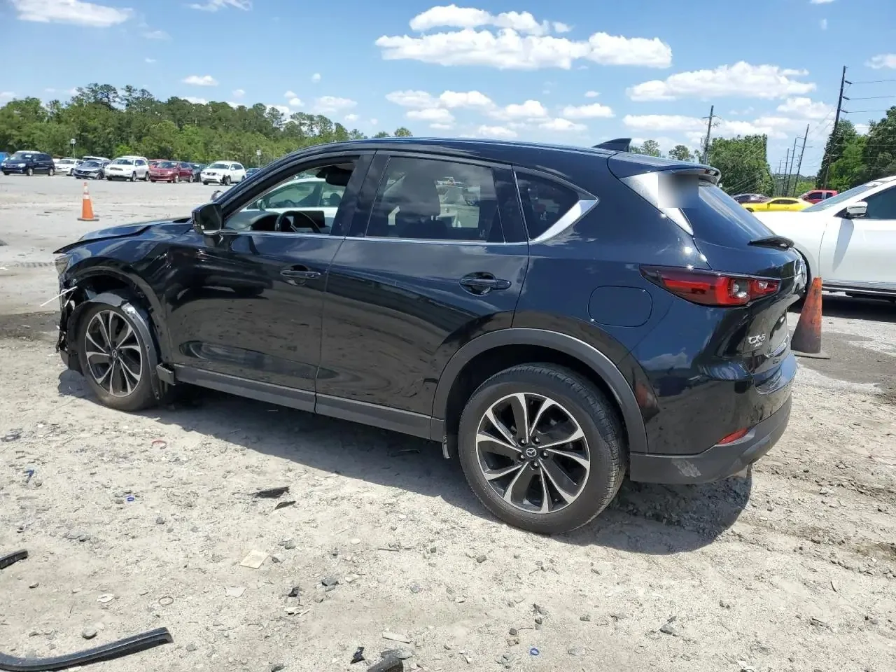 The damaged black CX-5 on the dirt auction lot from the rear left, with the front bumper torn open at the far corner and the rest of the body straight. Marker writing on the rear glass is blurred