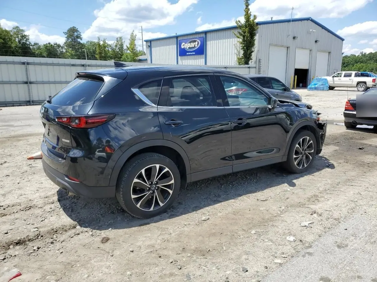 The black CX-5 from the rear right on the auction lot, with the torn front corner at the far end and a metal Copart building behind it. The plate on another car is blurred