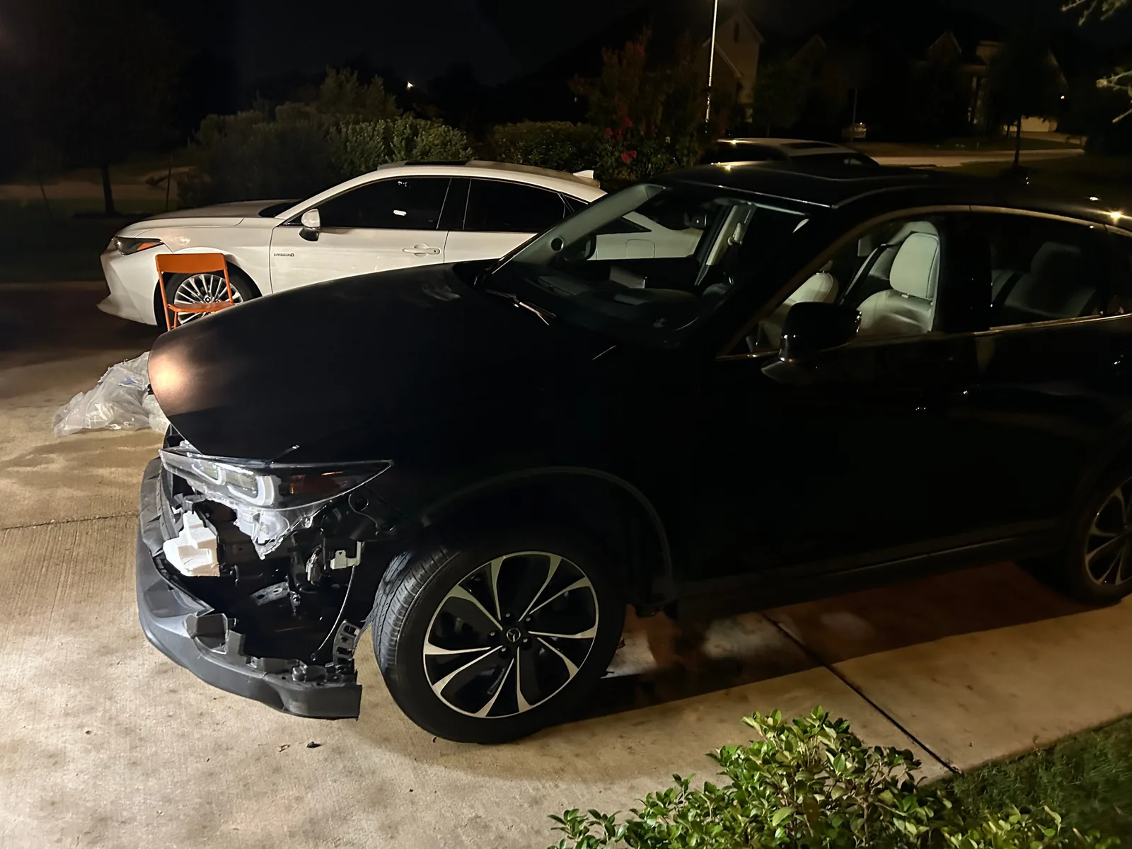 The CX-5 at night from the front left in a driveway, with the dull black hood and the headlight fitted, and the corner below the headlight open with parts and wiring showing. A white car is parked beside it and a street sign in the distance is blurred
