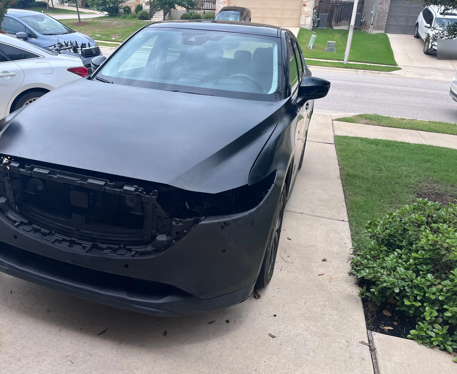 The CX-5 in daylight seen head on in a driveway, with a dull black hood and bumper cover and an empty grille frame where the grille goes. The plate on a car across the street is blurred